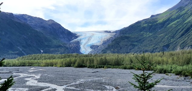 Glacier between two green mountains with riverbed in foreground.