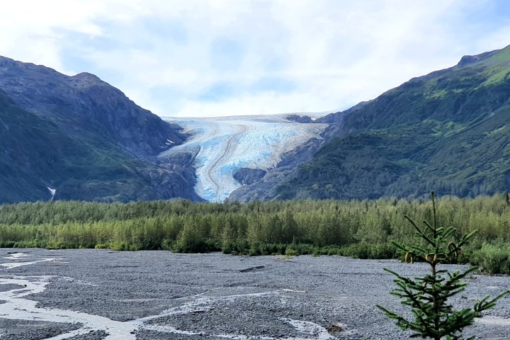 Glacier between two green mountains with riverbed in foreground.