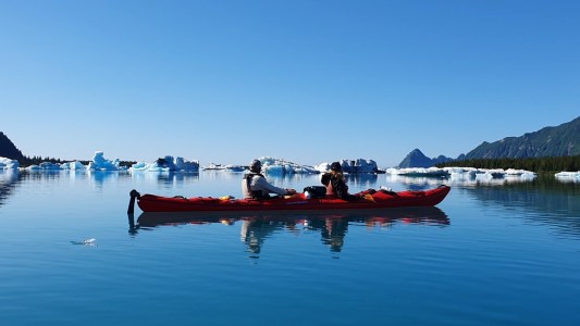 Two people kayaking on calm water with icebergs in the background.