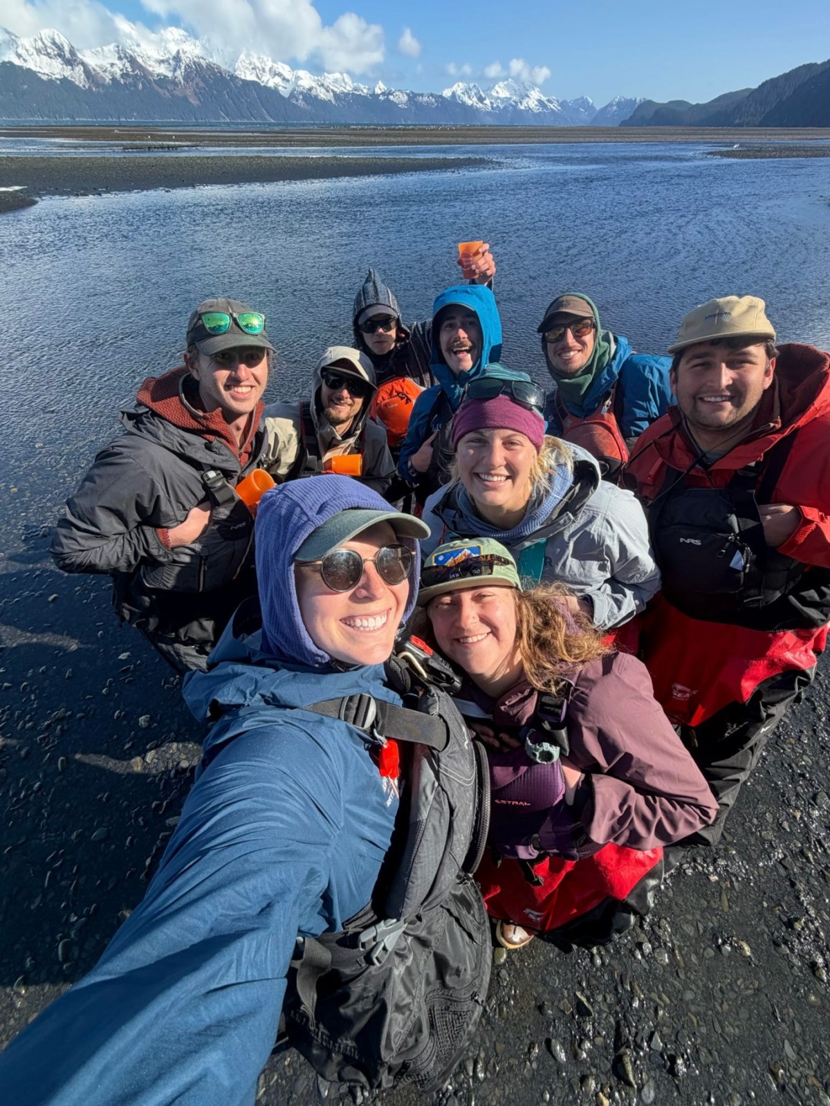 Group selfie outdoors by a lake with snow-capped mountains in the background.