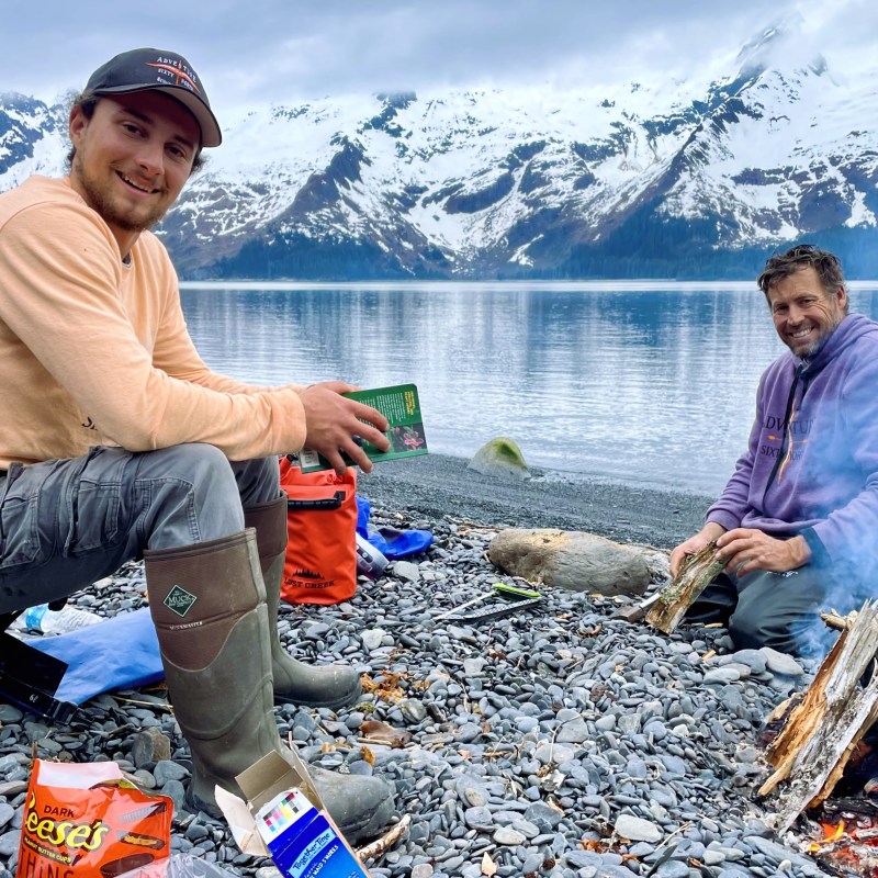 Two people sitting by a lakeside campfire with mountains in the background.