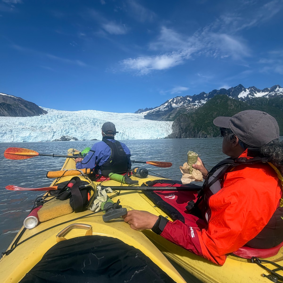 Two people kayaking near a glacier under a clear blue sky.